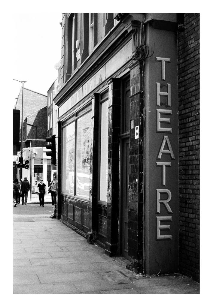 Black and white film photograph of the side of a building on the street that reads "Theatre" in big letters.