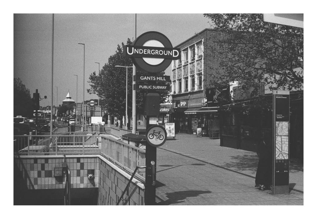 Black and white film photograph of a London underground sign that reads "Giants Hill" next to a street.