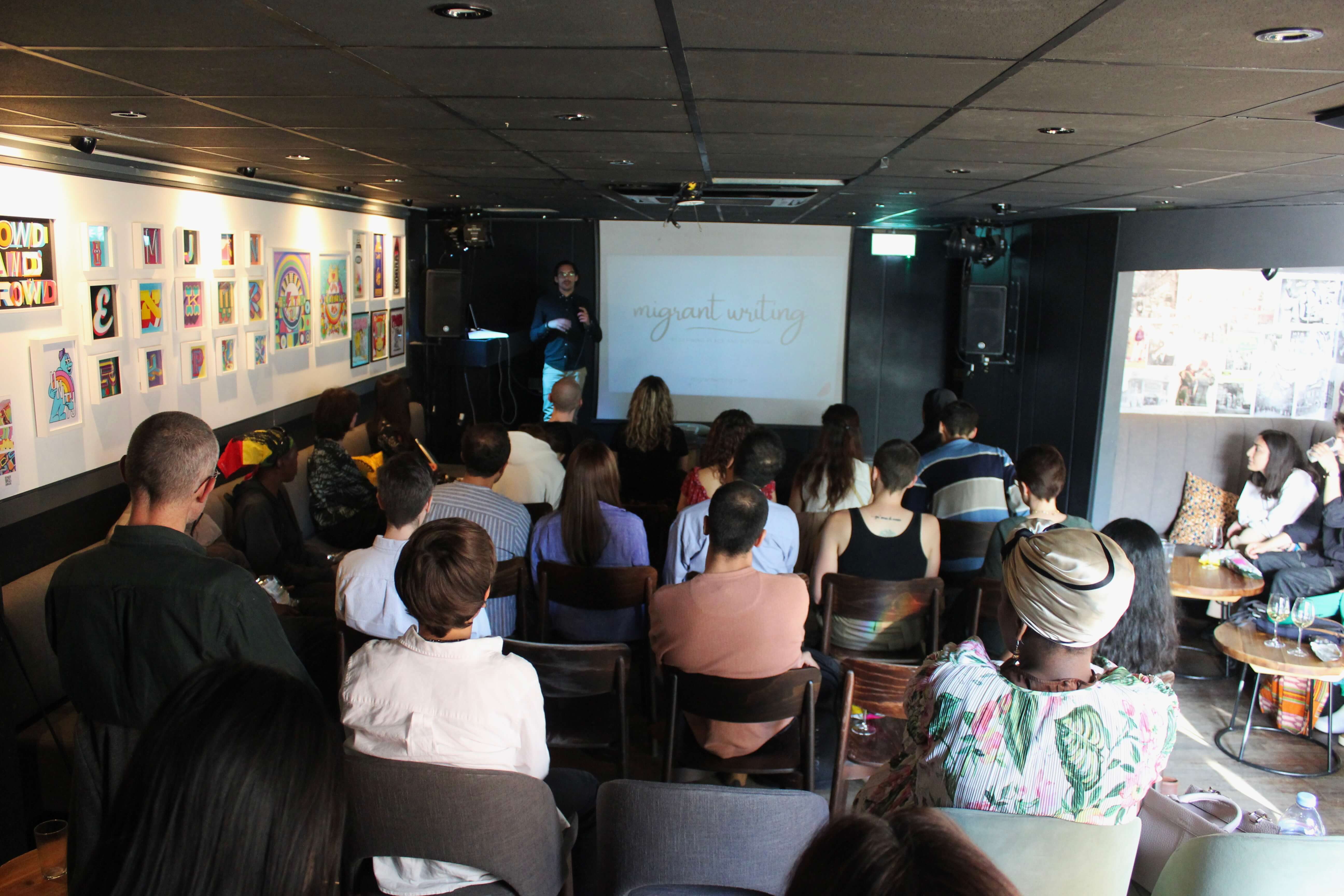 Gabriele Lazzari presenting the migrant project on stage with a view of the back of the audience.
